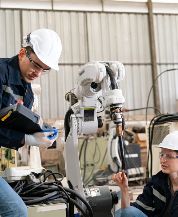 Two engineers in hard hats work with a robotic arm in a workshop. One engineer inspects a device while the other communicates, showcasing teamwork in automation.