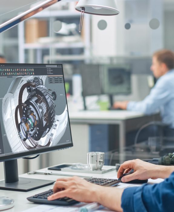 Over the Shoulder Shot of Engineer Working with CAD Software on Desktop Computer, Screen Shows Technical Details and Drawings. In the Background Engineering Facility Specialising on Industrial Design